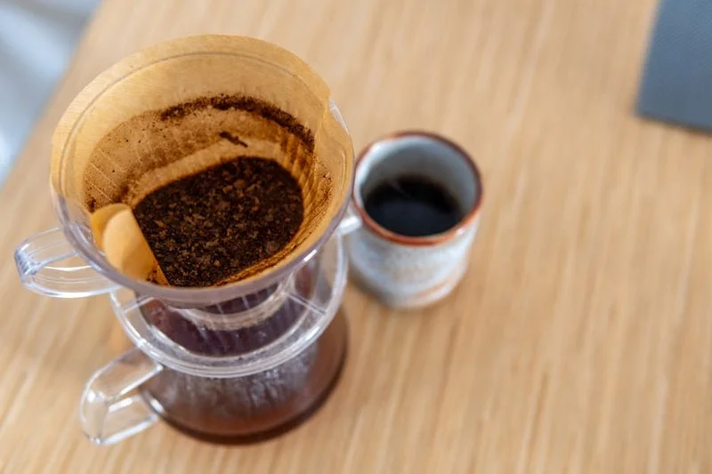 A barista preparing a pour-over specialty coffee, with a selection of small, delicate pastries on a wooden board
