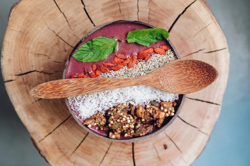 Lebendige Superfood Acai Bowl mit frischen Beeren, Granola, Kokosflocken und einem grünen Smoothie im Hintergrund auf einem Holztisch