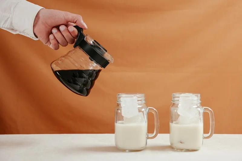 A person's hand carefully pouring dark cold brew coffee into a glass filled with ice and a splash of milk